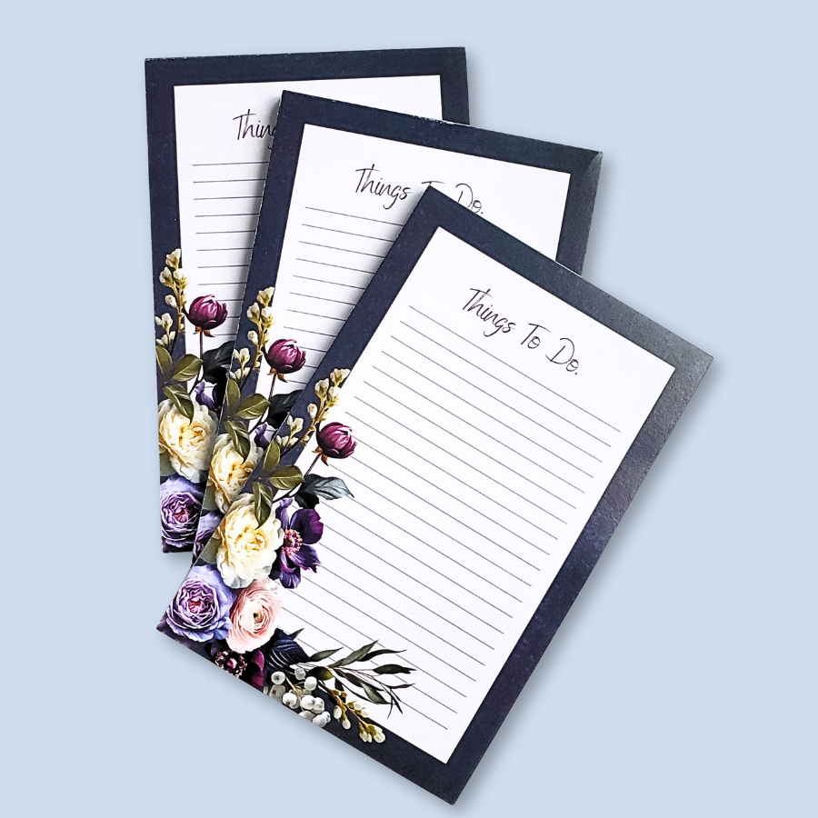 A vertical lined notepad with the heading 'Things To Do.' at the top, framed by a black border and decorated with purple, cream, and pink flowers along the lower left corner, shown on a light blue background.