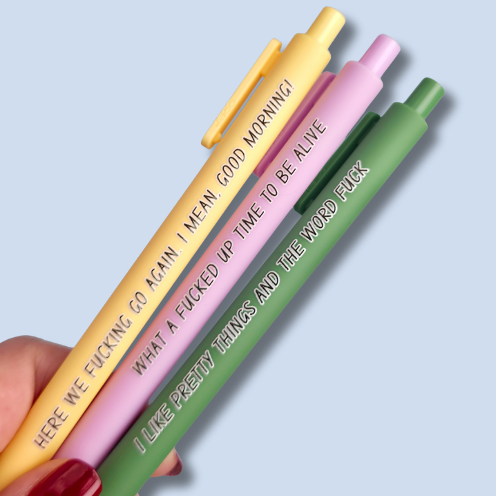Three pens with sayings on them. "here we fucking go again. I mean, Good morning!"(yellow pen), "What a fucked up time to be alive" (lilac pen), "I like pretty things and the word fuck" ( moss green pen)