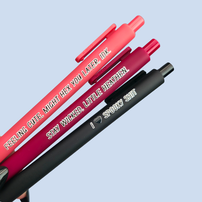 Three pens with humorous text on a light blue background. Text reads, "Feeling cute, might hex you later, IDK" on a salmon pink pen, "Stay wicked, little heathen" on a wine pen and "I  heart spooky shit" on a black pen