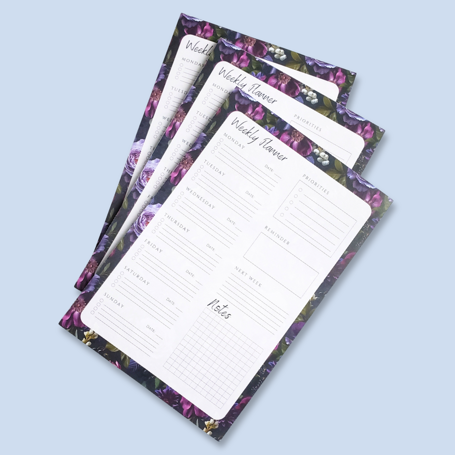 A weekly planner notepad with a white writing area featuring sections for Monday through Sunday, a priorities list, a reminder box, and a notes grid, surrounded by a purple and green floral border, shown on a light blue background.