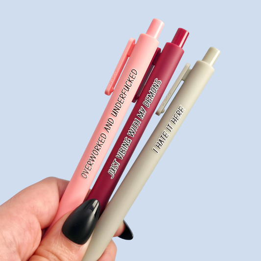 Three pens with humorous text on a light blue background. Text reads, "Overworked and underfucked" on a pastel pink pen, "Just vibing with my demons" on a dark red pen and "I hate it here" on a light grey pen