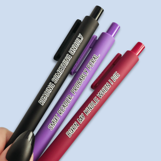 Three pens with quotes. "Reading something Unholy" on a black pen, "smut reader. Probably feral." on a purple pen, and "burn my Kindle when I die" on a red pen.