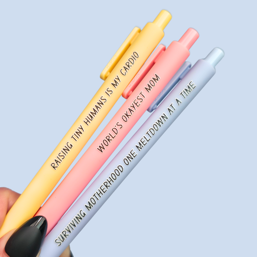 Three pens with humorous text on a light blue background. Text reads, "Raising tiny humans is my cardio" on a pastel yellow pen, "World's okayest mom" on a pastel pink pen and "surviving motherhood one meltdown at a time" on a pastel blue pen