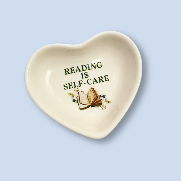 A heart-shaped ceramic dish in cream white with the text 'READING IS SELF-CARE' and an illustration of an open book and small flowers, shown on a light blue background.