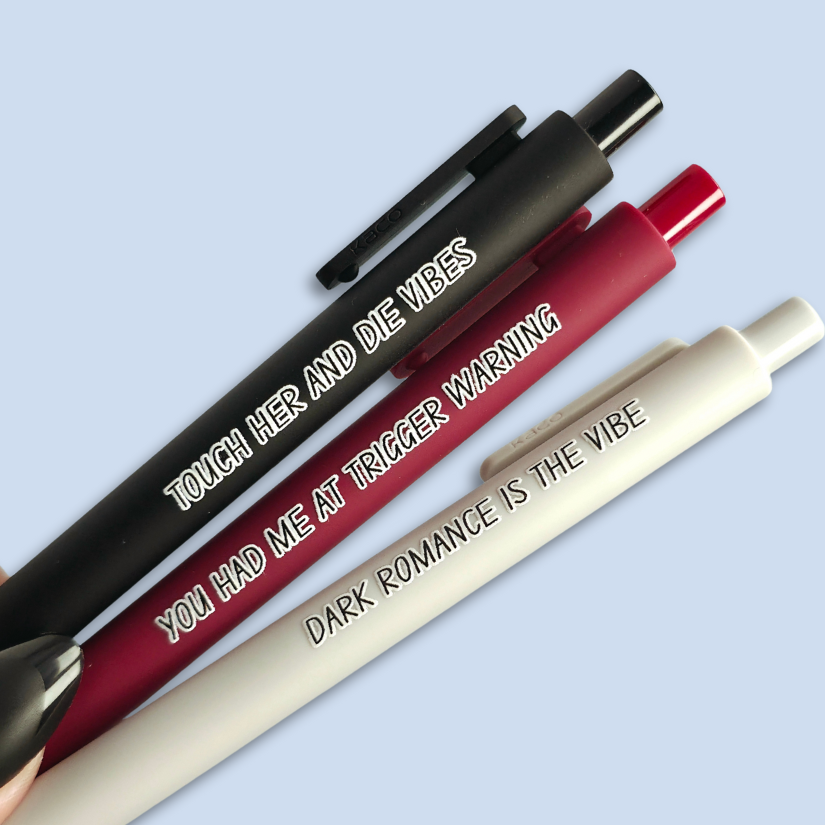 Three pens with sayings on them. "Dark romance is the vibe" (light grey pen), "You had me at trigger warning" (wine pen), "Touch her and die vibes" ( black pen).