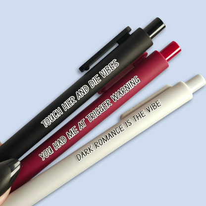 Three pens with sayings on them. "Dark romance is the vibe" (light grey pen), "You had me at trigger warning" (wine pen), "Touch her and die vibes" ( black pen).