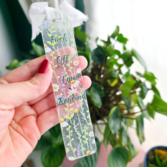A hand with red nail polish holds a clear, glittery rectangular bookmark featuring a floral design and the text 'Fuck Off I'm Reading'. The background shows a soft-focus indoor setting with green houseplants.