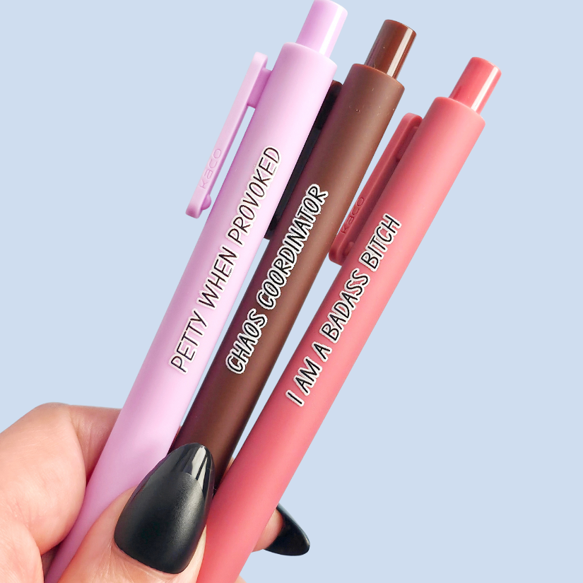 Three pens with humorous text. "Petty when provoked" on a lilac pen, "chaos coordinator" on a brown pen, and "I am a badass bitch" on a pink pen
