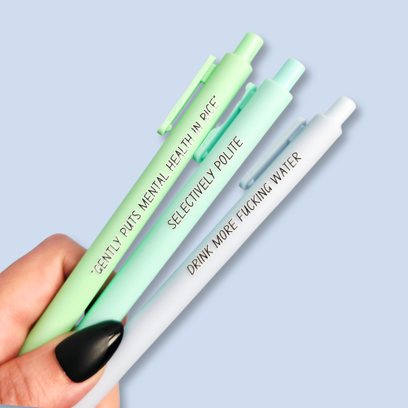Three pens with humorous text on a light blue background. Text reads, "Gently puts mental health in rice" on a pastel green pen, "Selectively polite" on a mint pen and "drink more fucking water" on a pastel blue pen