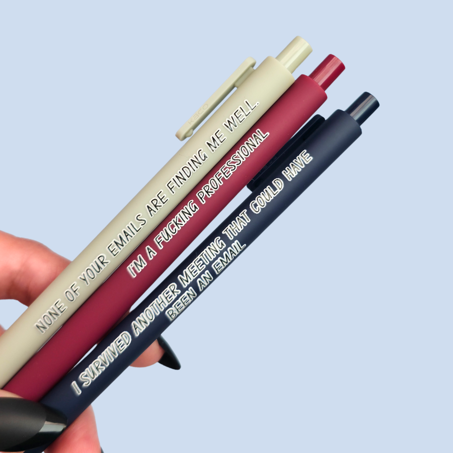 Three pens with quotes. "None of your emails are finding me well" on a grey pen, " I'm a fucking professional" on a dark red pen, and "I survived another meeting that could have been an email" on a navy blue pen.