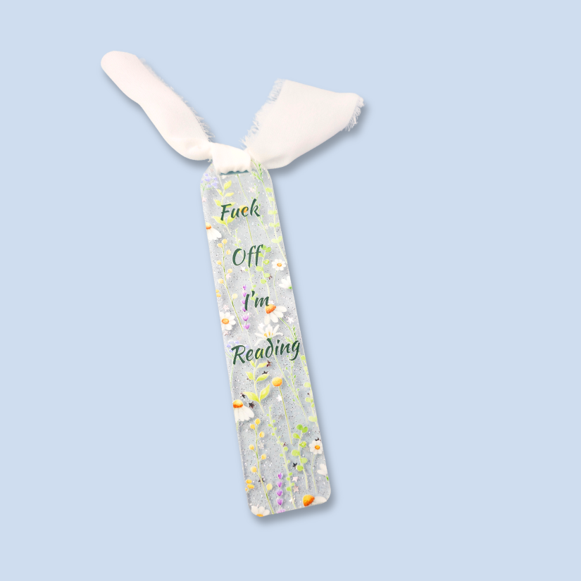 A clear rectangular bookmark with white tassels at the top, printed with the phrase 'Fuck Off I'm Reading' in green script and decorated with small white flowers and colorful wildflower illustrations, shown on a light blue background.