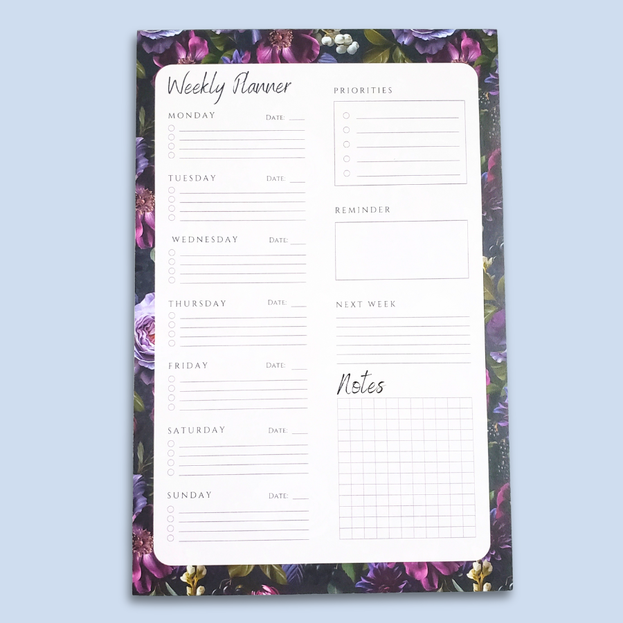 A weekly planner notepad with a white writing area featuring sections for Monday through Sunday, a priorities list, a reminder box, and a notes grid, surrounded by a purple and green floral border, shown on a light blue background.