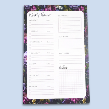 A weekly planner notepad with a white writing area featuring sections for Monday through Sunday, a priorities list, a reminder box, and a notes grid, surrounded by a purple and green floral border, shown on a light blue background.