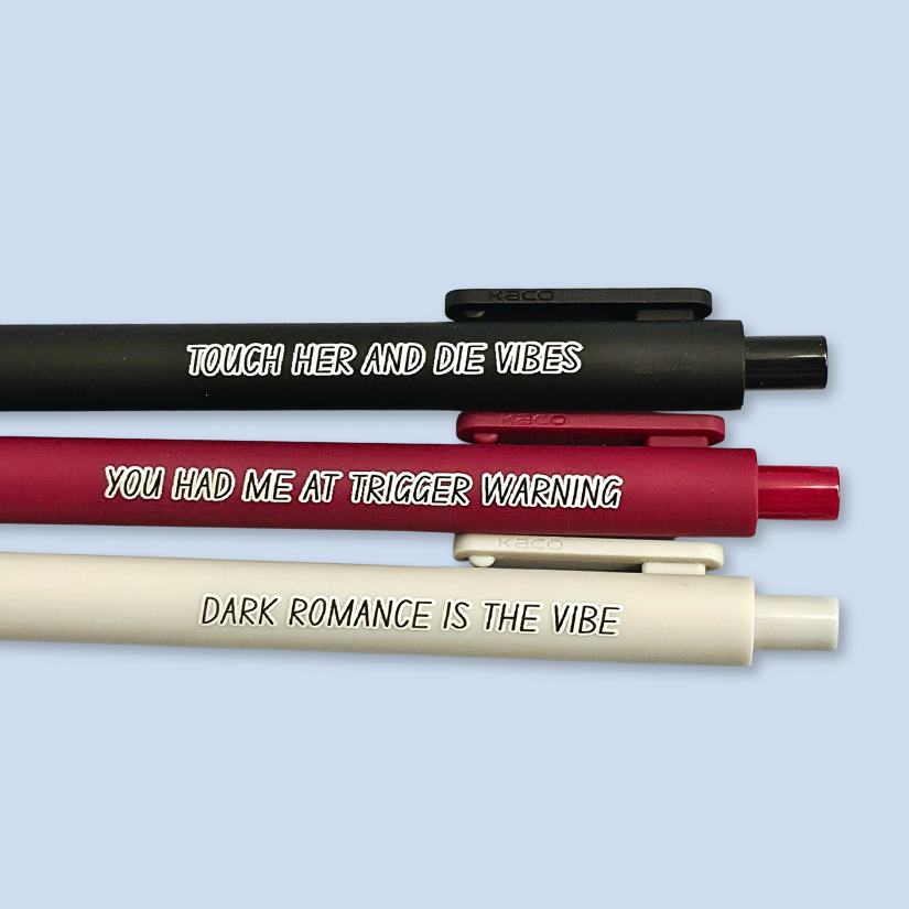 Three pens with sayings on them. "Dark romance is the vibe" (light grey pen), "You had me at trigger warning" (wine pen), "Touch her and die vibes" ( black pen).