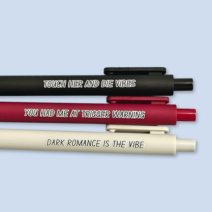 Three pens with sayings on them. "Dark romance is the vibe" (light grey pen), "You had me at trigger warning" (wine pen), "Touch her and die vibes" ( black pen).