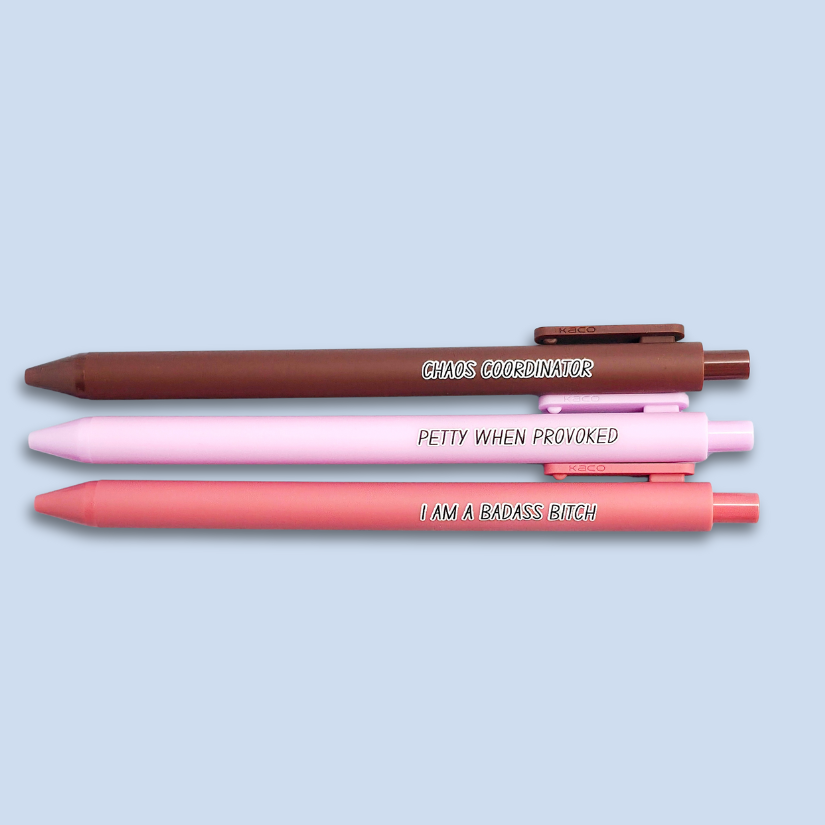 Three pens with humorous text. "Petty when provoked" on a lilac pen, "chaos coordinator" on a brown pen, and "I am a badass bitch" on a pink pen