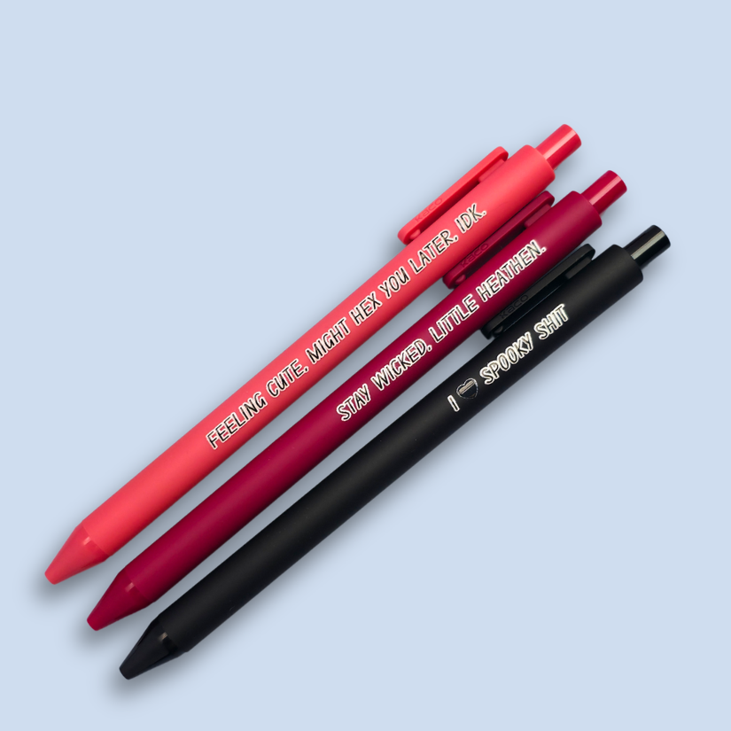 Three pens with humorous text on a light blue background. Text reads, "Feeling cute, might hex you later, IDK" on a salmon pink pen, "Stay wicked, little heathen" on a wine pen and "I  heart spooky shit" on a black pen