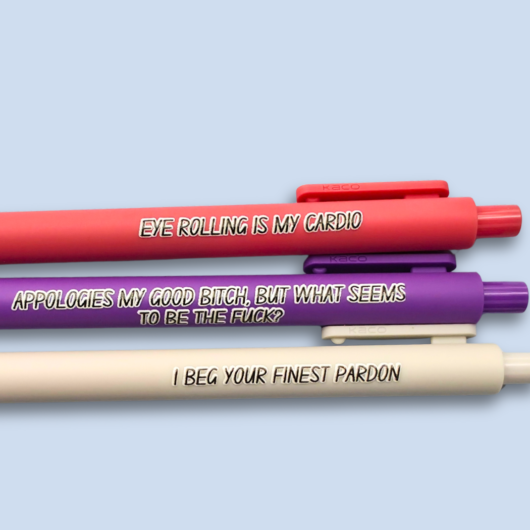 Three novelty gel pens in red, purple, and beige, stacked horizontally on a light blue background. Each pen features a humorous printed phrase in black with a white outline.