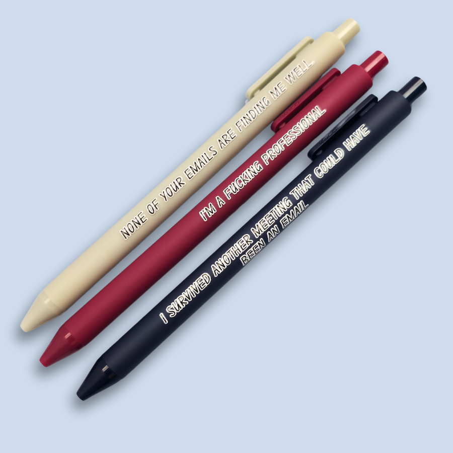 Three pens with quotes. "None of your emails are finding me well" on a grey pen, " I'm a fucking professional" on a dark red pen, and "I survived another meeting that could have been an email" on a navy blue pen.