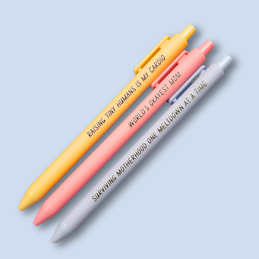 Three pens with humorous text on a light blue background. Text reads, "Raising tiny humans is my cardio" on a pastel yellow pen, "World's okayest mom" on a pastel pink pen and "surviving motherhood one meltdown at a time" on a pastel blue pen
