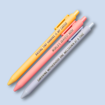 Three pens with humorous text on a light blue background. Text reads, "Raising tiny humans is my cardio" on a pastel yellow pen, "World's okayest mom" on a pastel pink pen and "surviving motherhood one meltdown at a time" on a pastel blue pen