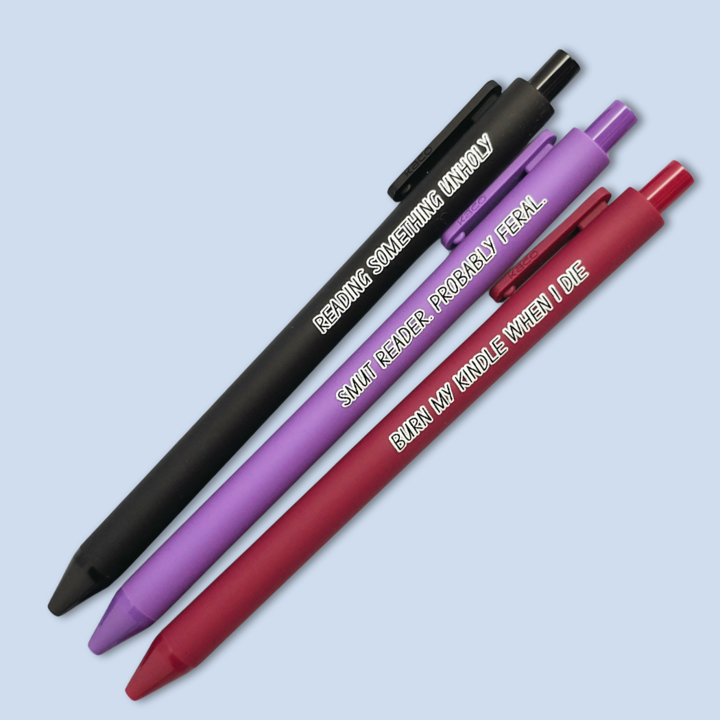 Three pens with quotes. "Reading something Unholy" on a black pen, "smut reader. Probably feral." on a purple pen, and "burn my Kindle when I die" on a red pen.