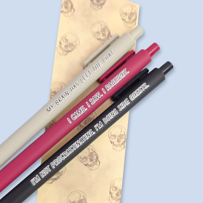 Three pens with humorous text on a skull-patterned bookmark and light blue background