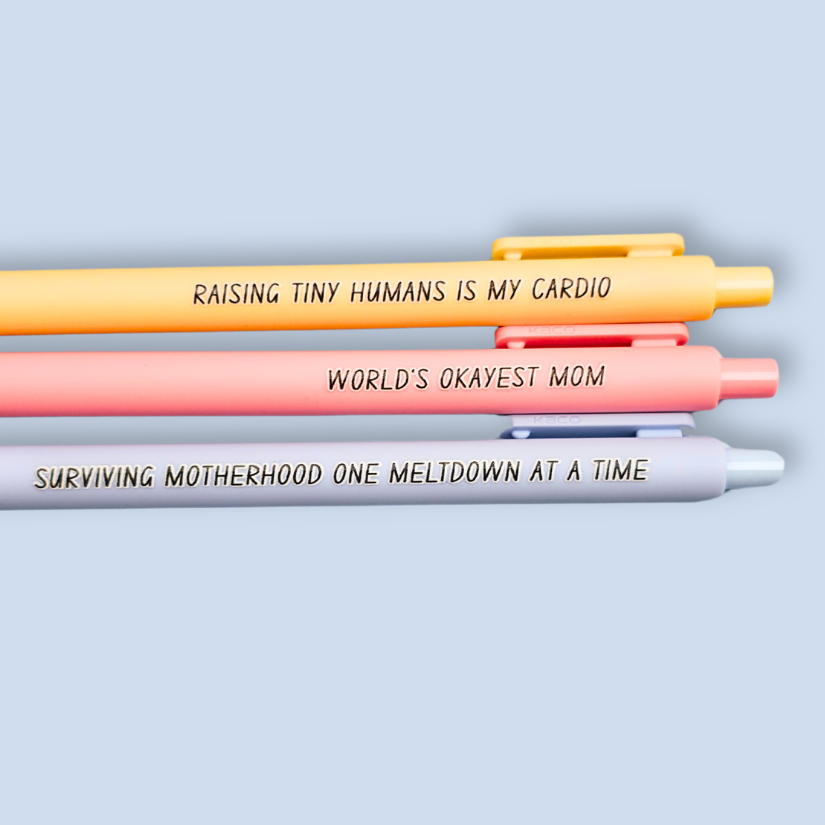 Three pens with humorous text on a light blue background. Text reads, "Raising tiny humans is my cardio" on a pastel yellow pen, "World's okayest mom" on a pastel pink pen and "surviving motherhood one meltdown at a time" on a pastel blue pen