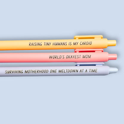 Three pens with humorous text on a light blue background. Text reads, "Raising tiny humans is my cardio" on a pastel yellow pen, "World's okayest mom" on a pastel pink pen and "surviving motherhood one meltdown at a time" on a pastel blue pen