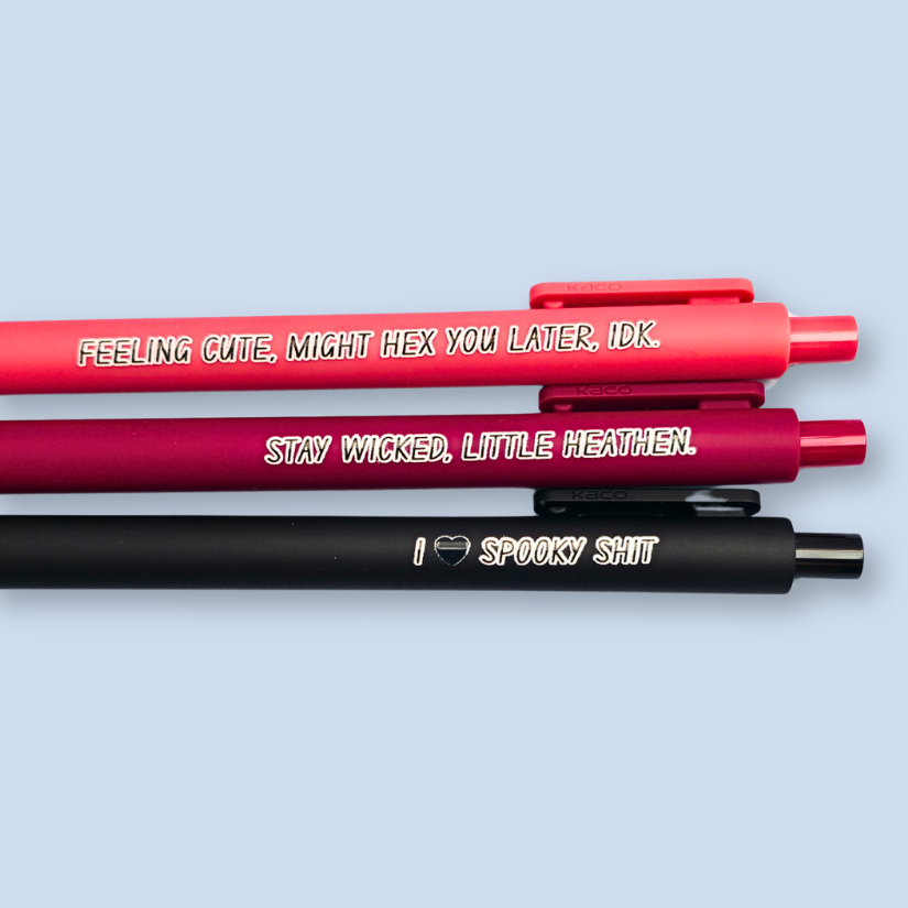 Three pens with humorous text on a light blue background. Text reads, "Feeling cute, might hex you later, IDK" on a salmon pink pen, "Stay wicked, little heathen" on a wine pen and "I  heart spooky shit" on a black pen