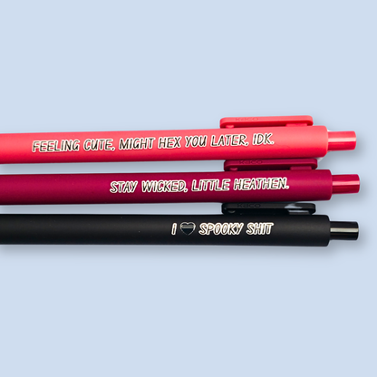 Three pens with humorous text on a light blue background. Text reads, "Feeling cute, might hex you later, IDK" on a salmon pink pen, "Stay wicked, little heathen" on a wine pen and "I  heart spooky shit" on a black pen