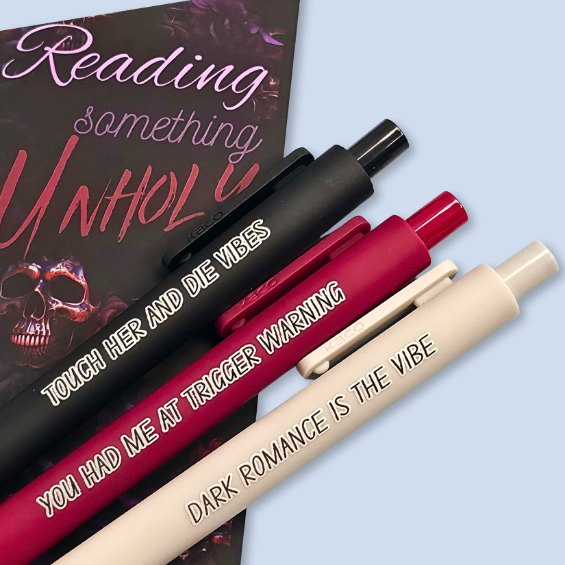 Three pens with sayings on them. "Dark romance is the vibe" (light grey pen), "You had me at trigger warning" (wine pen), "Touch her and die vibes" ( black pen).