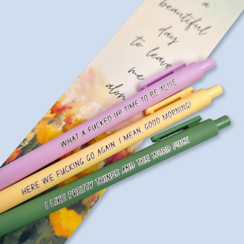 Three pens with sayings on them. "here we fucking go again. I mean, Good morning!"(yellow pen), "What a fucked up time to be alive" (lilac pen), "I like pretty things and the word fuck" ( moss green pen) Bookmark on the background