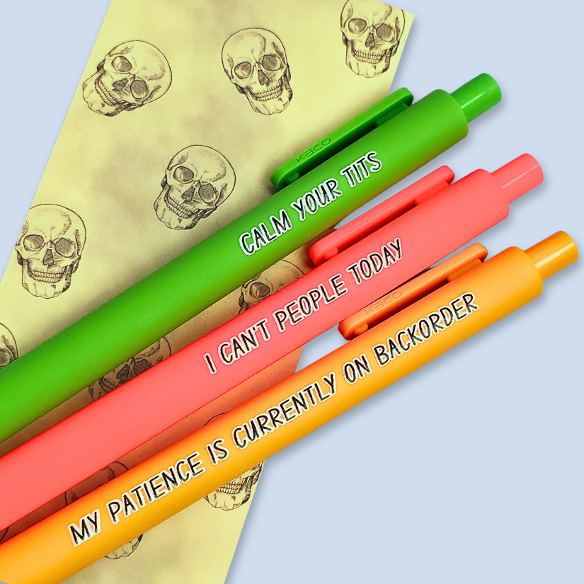 Three colorful pens with humorous text on a light blue background over a yellow bookmark with skull designs.