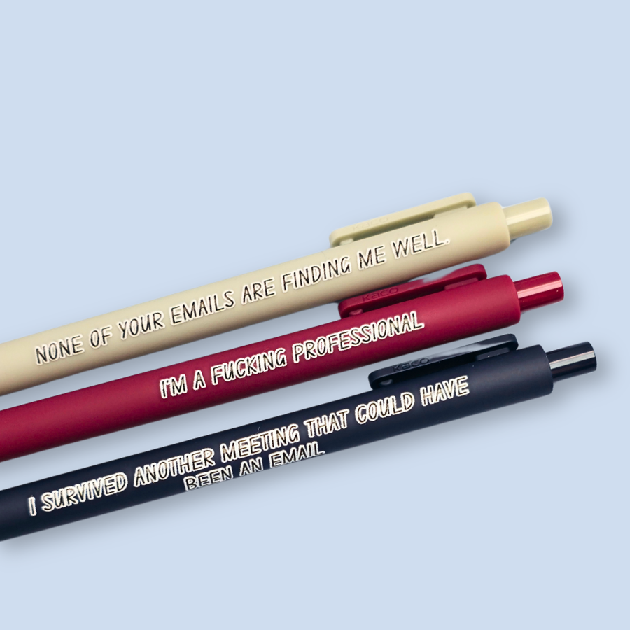 Three pens with quotes. "None of your emails are finding me well" on a grey pen, " I'm a fucking professional" on a dark red pen, and "I survived another meeting that could have been an email" on a navy blue pen.