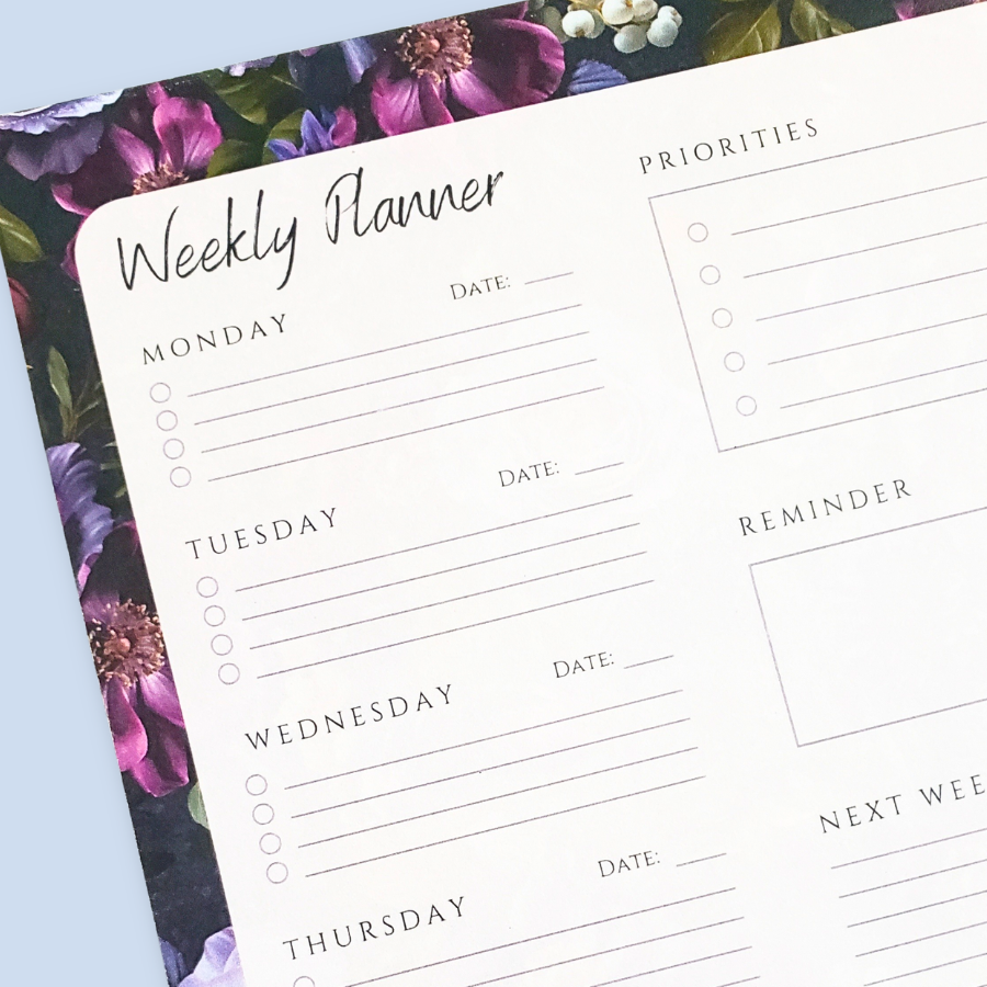 A weekly planner notepad with a white writing area featuring sections for Monday through Sunday, a priorities list, a reminder box, and a notes grid, surrounded by a purple and green floral border, shown on a light blue background.