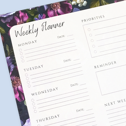 A weekly planner notepad with a white writing area featuring sections for Monday through Sunday, a priorities list, a reminder box, and a notes grid, surrounded by a purple and green floral border, shown on a light blue background.