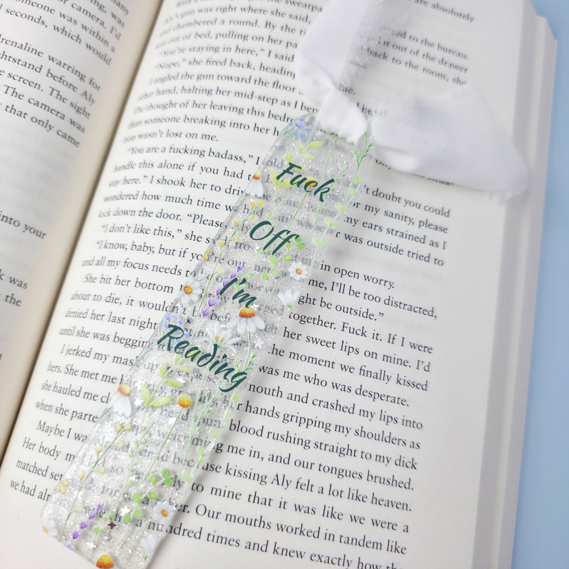 An open book with printed text, overlaid with a decorative floral bookmark or ribbon featuring purple flowers, green leaves, and the words 'Fuck Off I'm Reading' in stylized lettering.