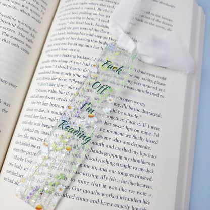 An open book with printed text, overlaid with a decorative floral bookmark or ribbon featuring purple flowers, green leaves, and the words 'Fuck Off I'm Reading' in stylized lettering.