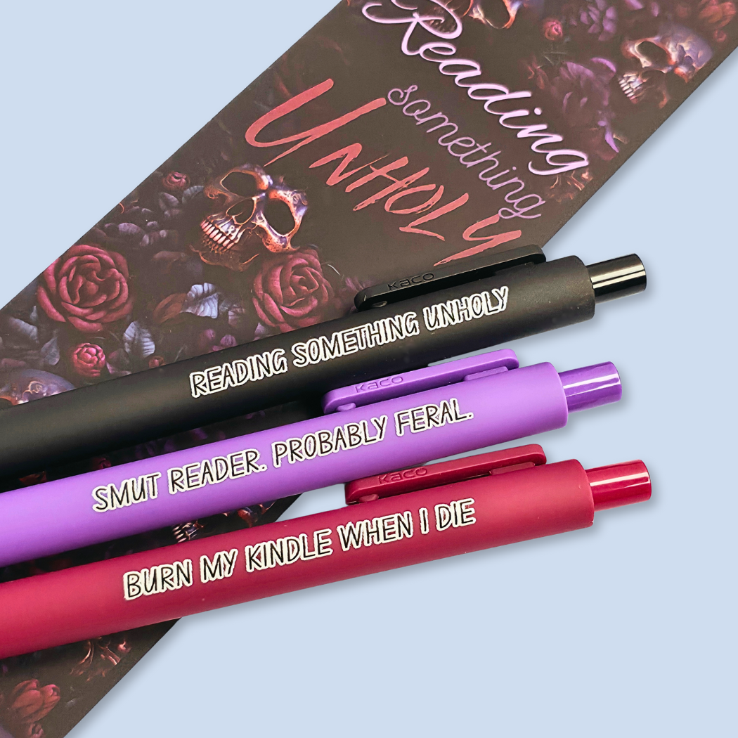 Three pens with quotes. "Reading something Unholy" on a black pen, "smut reader. Probably feral." on a purple pen, and "burn my Kindle when I die" on a red pen.