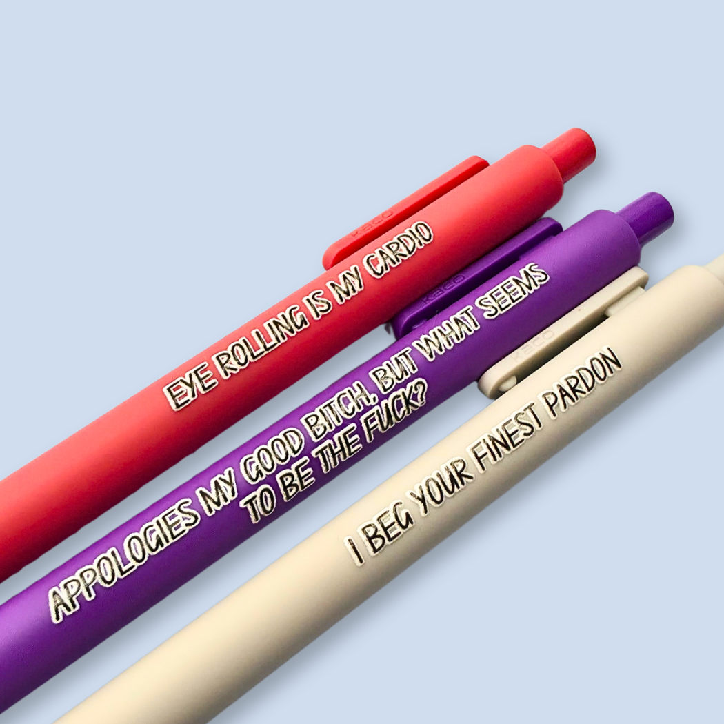 Three novelty ballpoint pens in red, purple, and beige, arranged diagonally on a light blue background. Each pen features humorous text printed on its barrel: 'EYE ROLLING IS MY CARDIO', 'APPOLOGIES MY GOOD BITCH, BUT WHAT SEEMS TO BE THE FUCK?', and 'I Beg YOUR FINEST PARDON'.