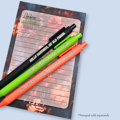 Three pens with quotes. "Hello darkness, my old friend." on a black pen, ' That's a terrible idea. What time?" on a lime green pen, and "I take super hot showers to practice burning in hell" on an orange pen. The pens rest on a notepad, with a sample ot the ink colors. Notepads are sold separately