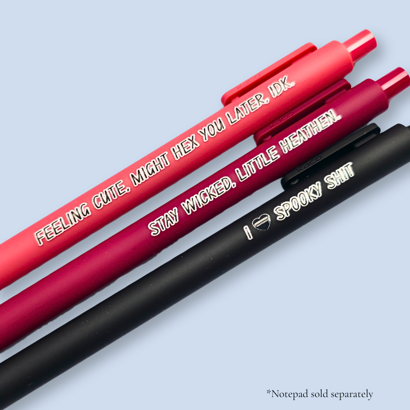 Three pens with humorous text on a light blue background. Text reads, "Feeling cute, might hex you later, IDK" on a salmon pink pen, "Stay wicked, little heathen" on a wine pen and "I  heart spooky shit" on a black pen