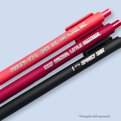 Three pens with humorous text on a light blue background. Text reads, "Feeling cute, might hex you later, IDK" on a salmon pink pen, "Stay wicked, little heathen" on a wine pen and "I  heart spooky shit" on a black pen