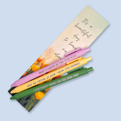 Three pens with sayings on them. "here we fucking go again. I mean, Good morning!"(yellow pen), "What a fucked up time to be alive" (lilac pen), "I like pretty things and the word fuck" ( moss green pen) Bookmark on the background