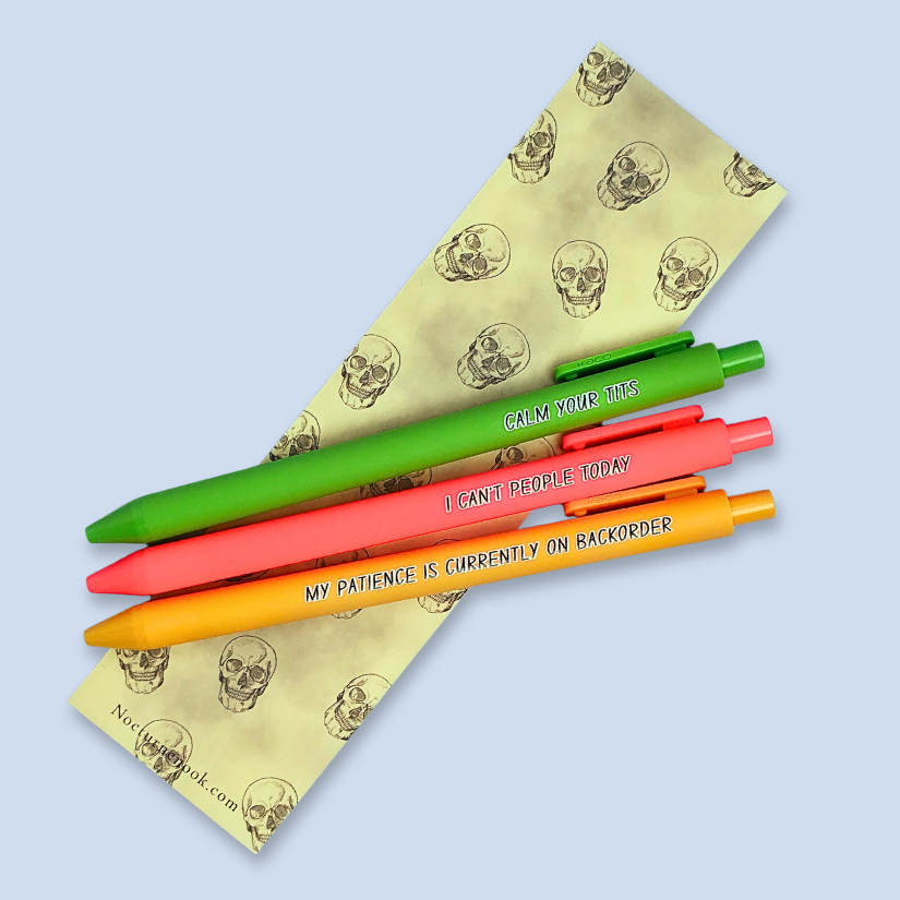 Three colorful pens with humorous text on a light blue background over a yellow bookmark with skull designs.