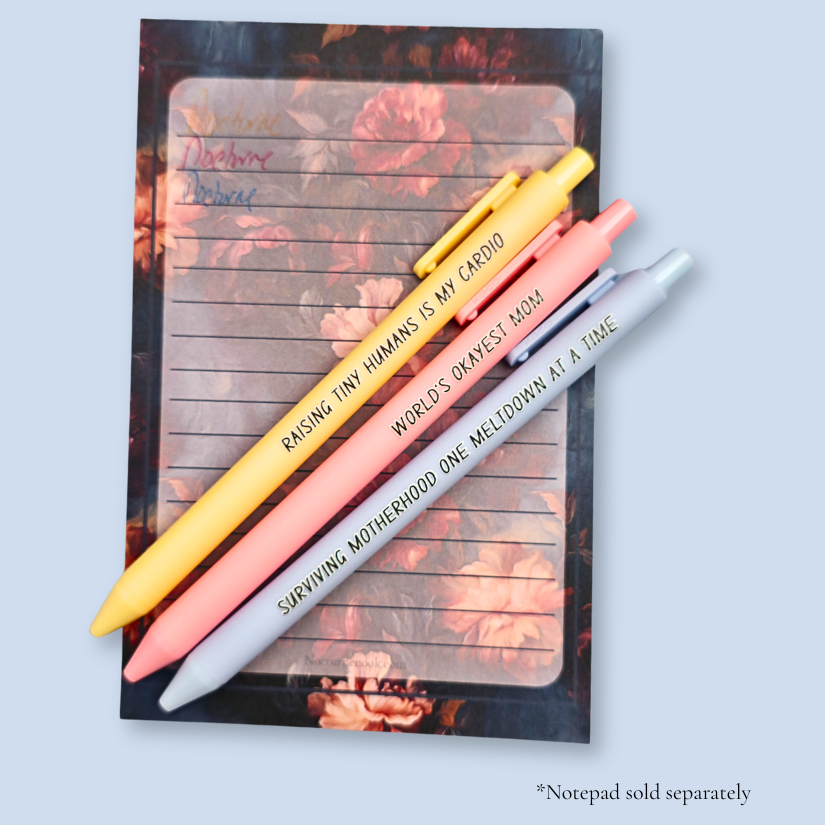 Three pens with humorous text on a light blue background. Text reads, "Raising tiny humans is my cardio" on a pastel yellow pen, "World's okayest mom" on a pastel pink pen and "surviving motherhood one meltdown at a time" on a pastel blue pen. Ink matches the color of the pen