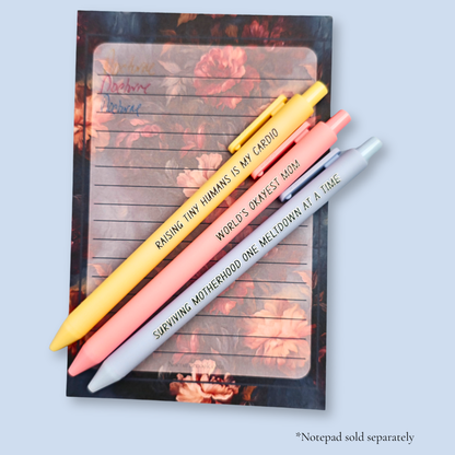 Three pens with humorous text on a light blue background. Text reads, "Raising tiny humans is my cardio" on a pastel yellow pen, "World's okayest mom" on a pastel pink pen and "surviving motherhood one meltdown at a time" on a pastel blue pen. Ink matches the color of the pen