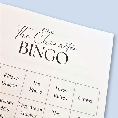 A close-up of a printed bingo card titled 'Find The Character Bingo' on a light blue background. The card has black text on white paper and shows some grid categories such as 'Rides a Dragon', 'Fae Prince', 'Loves Knives', and 'Growls'.