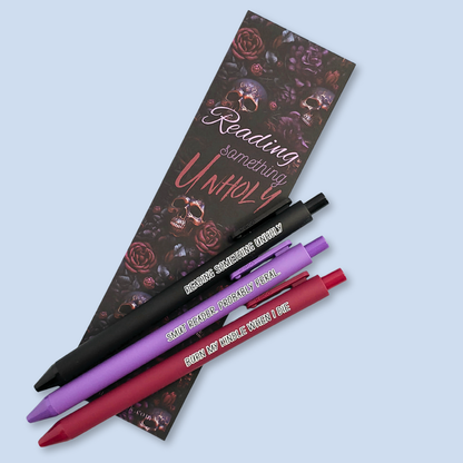 Three pens with quotes. "Reading something Unholy" on a black pen, "smut reader. Probably feral." on a purple pen, and "burn my Kindle when I die" on a red pen. Floral and skulls bookmark in the background.